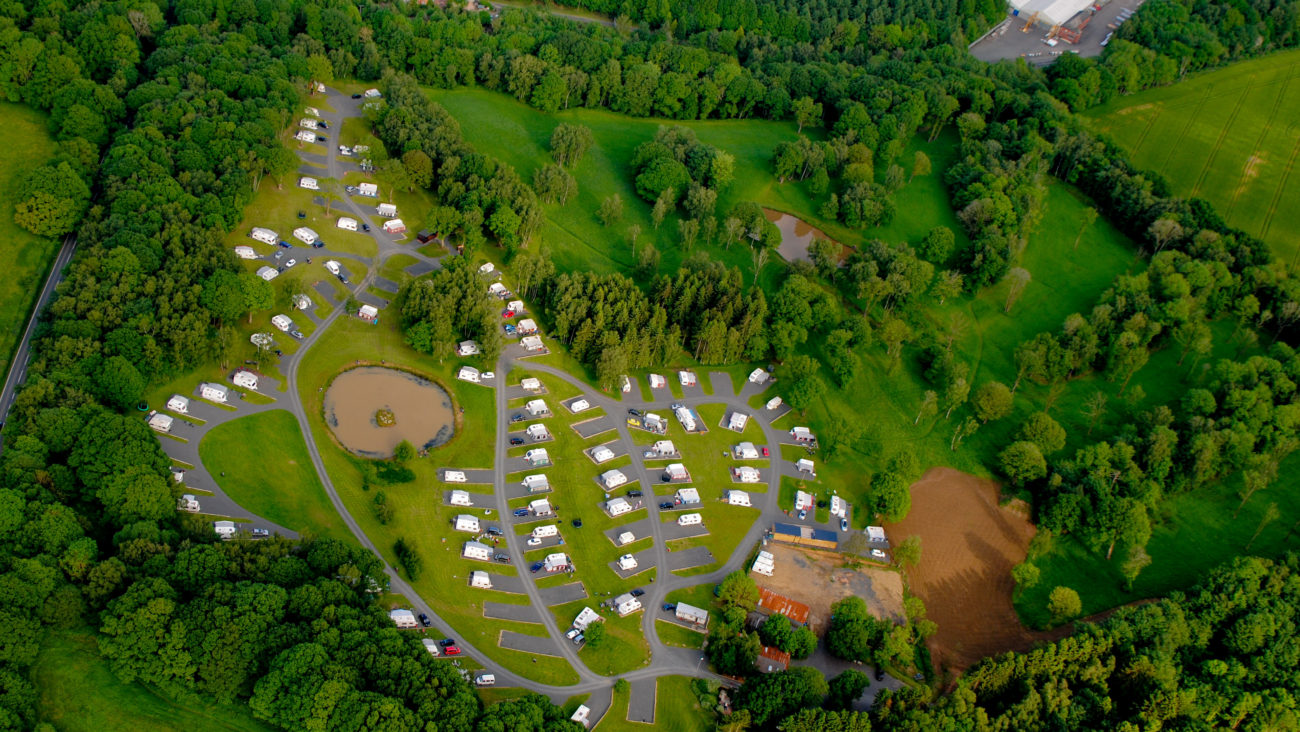 An aerial photograph of Lodge Coppice. Well spaced caravans, trees and the pool all seen from above.